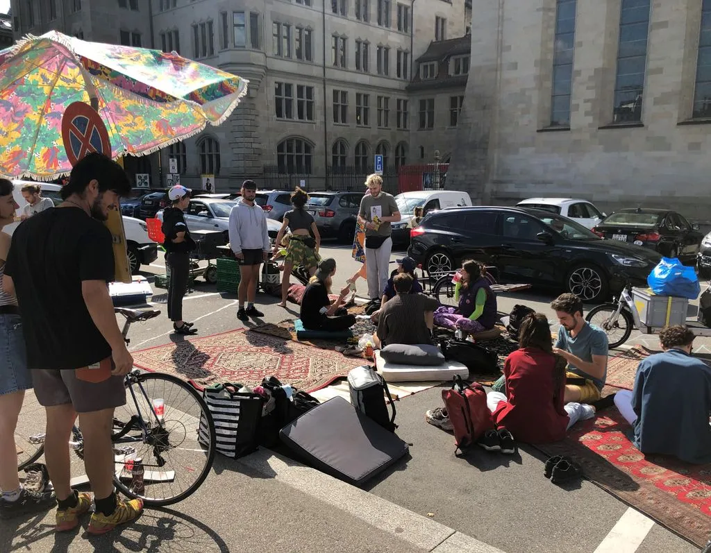 Eine Gruppe von jungen Menschen sitzt auf Teppichen auf einem öffentlichen Parkplatz.