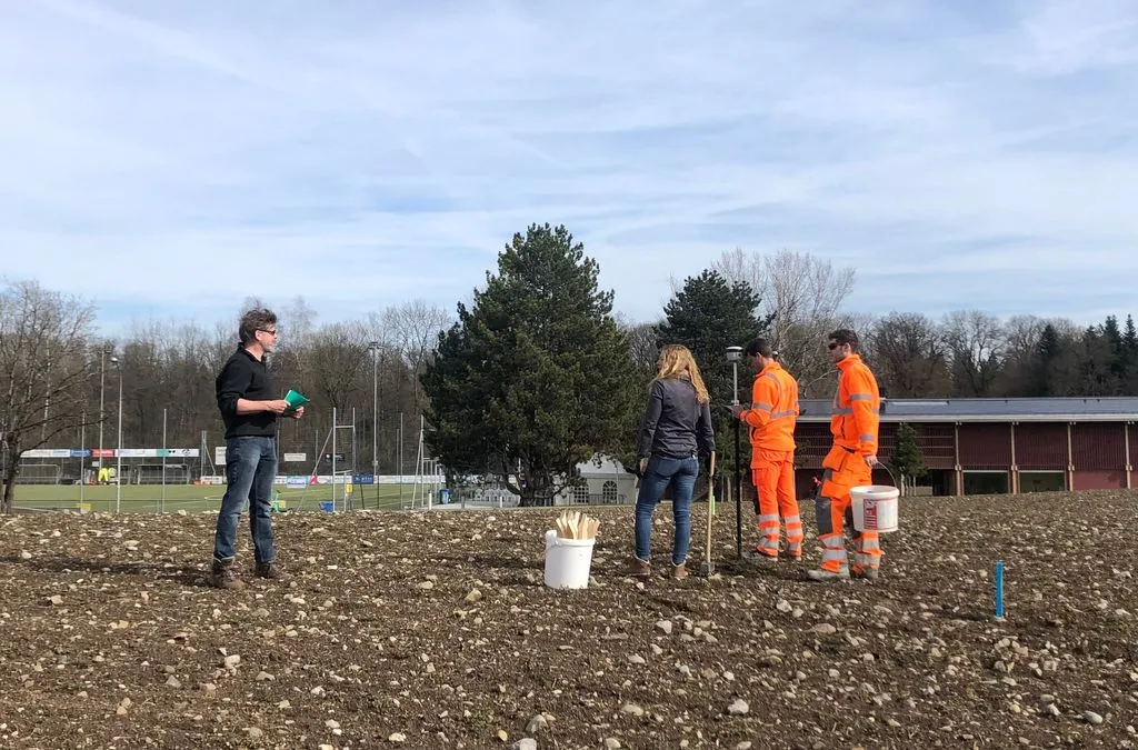 Vier Personen vermessen ein braches Stück Land. Zwei tragen orange Leuchtkleidung.
