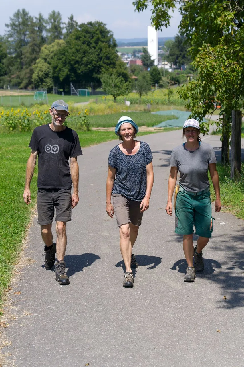 Die drei Pächter:innen des Huebhofs gehen auf einem sonnigen Feldweg.