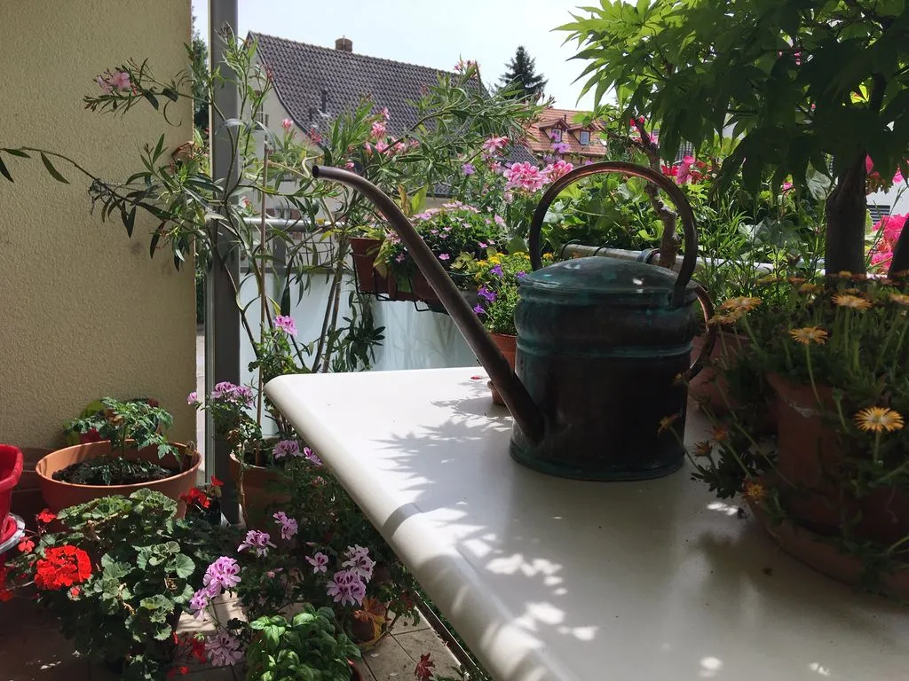 Eine kleine kunstvolle Giesskanne steht auf einem Balkon mit Blumen.
