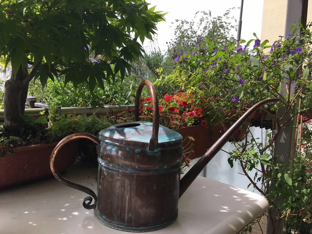 Eine kleine kunstvolle Giesskanne steht auf einem Balkon mit Blumen.