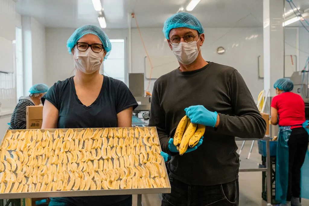 Zwei Personen mit Maske, Handschuhen und Haarnetz halten ein Blech mit geschnittenen Bananen.